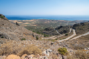 Serpentine road in barren rocky hillside with sea in distance in sunshine with clear blue sky, Greece, island of Crete, wide angle lens