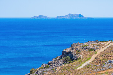 Barren rocky mountainside on a beach with calm sea, two islands in the distance, sunshine, telephoto