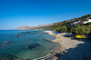Sunny sandy seaside beach with clear transparent water, blue sky, houses, umbrellas and sunbeds, Plakias, Greece, Crete island, wide angle lens