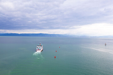 A ship sails smoothly on a vast green lake, surrounded by calm waters and red buoys. The cloudy sky and distant mountain range create a peaceful atmosphere of a summer day.