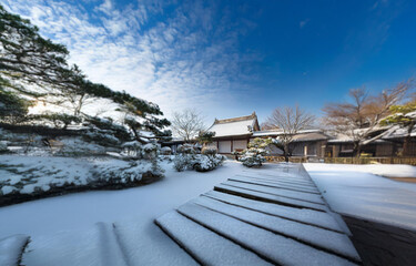 A snow covered wooden path leads to a traditional building in a Japanese garden with a clear blue sky and fluffy white clouds.