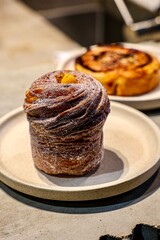 Sugar-coated cruffin on display at a bakery. A close-up shot of a beautifully crafted cruffin dusted with sugar, displayed on a plate. In the background, another pastry adds depth to the scene.