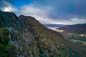 Forested hills and cliffs with distant lake under dramatic skies. A stunning view of forested hills and steep rocky cliffs, with a tranquil lake visible in the distance.