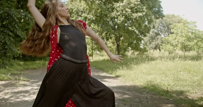 Woman in red and black outfit performs an interpretive modern dance routine in a sunny park.