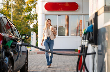 Young blonde attractive woman on gas station going towards the car with coffee while hose is in the tank 