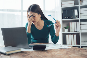 Obraz premium Overwhelmed Businesswoman: A stressed young woman sits at her desk, rubbing her temples in exhaustion, surrounded by paperwork and a laptop. The image conveys the pressures of modern work life.