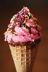 Close-up of a delicious pink ice cream cone with colorful sprinkles and chocolate sauce on a waffle cone, perfect for summer treats.