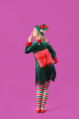 Beautiful young woman dressed as elf with Christmas gift showing silence gesture on purple...
