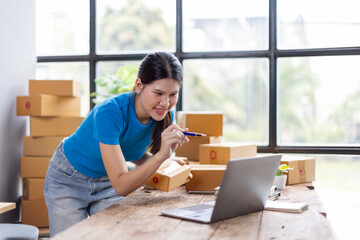 Asian young female sme business owner with product package box at warehouse, merchant seller checking customer address order confirming parcel delivery.
