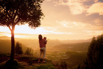 A woman is peacefully standing beneath a tall, lush tree, gazing at the beautiful sunset as the...