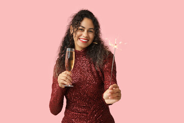 Happy African-American woman with Christmas sparkler and champagne on pink background