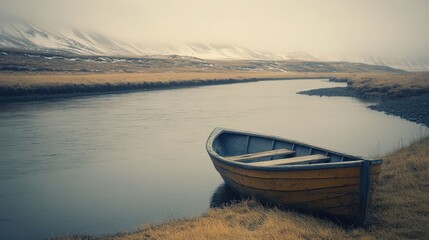A wooden boat resting near a calm river, surrounded by grassland and snowy mountains on a foggy, overcast day.
