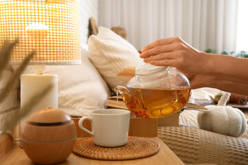 Female hands pouring green tea from teapot into cup near diffuser and lamp on bedside table in bedroom. Closeup