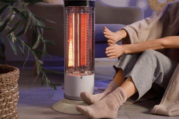 Young woman with blanket warming hands near electric heater on floor in evening at home, closeup