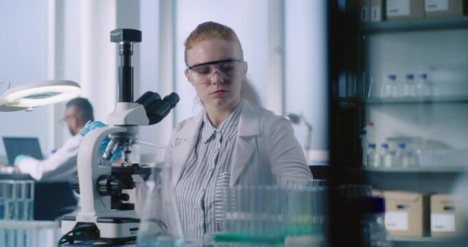 Bright medical science laboratory: Female biochemist takes liquid from test tube, looks under microscope, does analysis of sample. Scientist, microbiologist develops drugs using high-tech equipment.