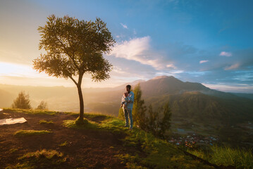 A man is confidently standing atop a picturesque hill, next to a lush tree that complements the...