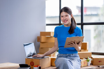 Smiling asian entrepreneur SME freelance sme business owner holding clipboard and working in packaging startup at home office, checking customer address order.