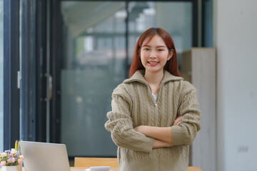 Confident young businesswoman keeping her arms crossed and looking at the camera while standing at her workspace.