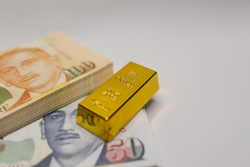 Shiny gold bar placed next to stacked Singapore dollar banknotes, representing wealth, finance, and investment opportunities in gold and currency.