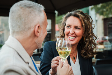 Smiling mature woman toasting with a white wine glass, enjoying a joyful moment with her husband at an outdoor restaurant, celebrating and sharing a special occasion with drinks and good company. - Powered by Adobe