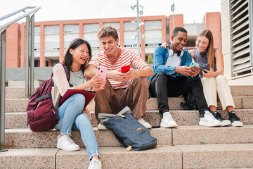 Group of university students sitting on outdoor stairs, laughing and using smartphones together, enjoying social media and online interaction during a break from their studies at campus.