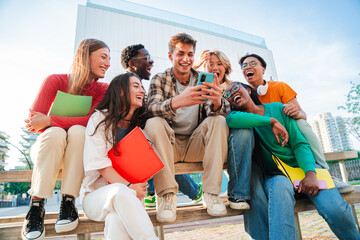 Group of diverse students sitting outdoors, laughing and using a cellphone together, enjoying social media, connecting through technology while holding notebooks and sharing moments on campus.