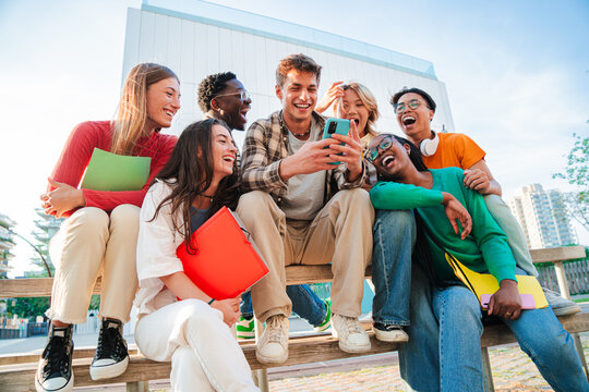 Group of diverse students sitting outdoors, laughing and using a cellphone together, enjoying social media, connecting through technology while holding notebooks and sharing moments on campus.