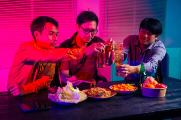 Three men are sitting at a table, holding glasses of beer and clinking glasses in celebration.They are enjoying food and drinks,eating and having fun in a party room with a happy,cheerful atmosphere