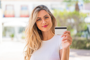 Pretty blonde Uruguayan woman holding a credit card at outdoors smiling a lot