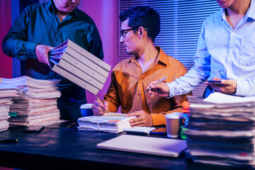 Three men work overtime at night, stressed and overloaded with tasks. They are exhausted, checking...