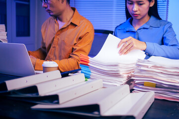 Obraz premium A businessman and woman are working overtime at their desks in office at night.They are busy, stressed, overloaded with too much work, checking documents and working on laptops while feeling tired