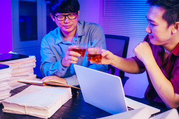 Two men are working late at the office, drinking beer while checking documents. They are overwhelmed with a lot of work, tired, and stressed as they continue to work through the night.