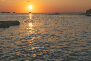 The sun sets over Cala Conta beach in Ibiza, Spain, casting a golden glow across the horizon. The calm waters reflect the warm light, with rocky formations adding depth to the serene coastal landscape