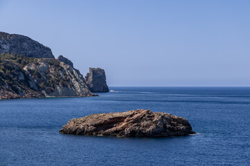 Ibiza rocky shores feature dramatic cliffs and a small islet surrounded by the azure waters of the Mediterranean. A boat glides through the calm sea, framed by the clear blue sky