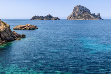 Picturesque seascape featuring the iconic Es Vedra and Es Vedranell islands near Ibiza, with a sailboat navigating the calm azure waters, framed by rocky cliffs and clear skies