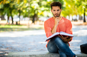 Fototapeta premium Handsome young man reading book on bench in the park