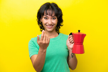 Obraz premium Young Argentinian woman holding coffee pot isolated on yellow background inviting to come with hand. Happy that you came