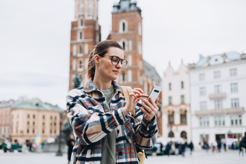 Fototapeta premium Beautiful stylish woman in glasses walking on Market Square in Krakow on autumn day and holding mobile phone. Phone Communication. Urban lifestyle concept. Check social networks, send sms.