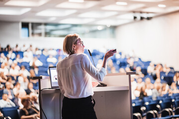 Female speaker giving a talk on corporate business conference. Unrecognizable people in audience at...