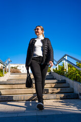 Beautiful middle-aged blonde woman wearing black elegant jacket, black jeans, cream turtleneck, handbag and sunglasses walking down concrete stairs in city on autumn day. Urban life. Woman in city.