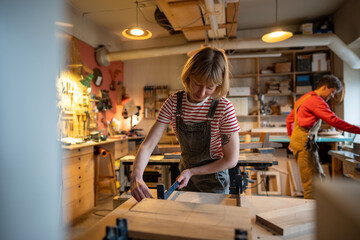 Female carpenter prepares wooden blocks for gluing, set up clamp in woodworking shop. Man and woman...