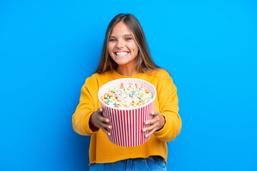 Young caucasian woman isolated on blue background holding a big bucket of popcorns