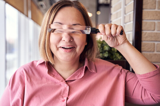 Fototapeta Woman enjoying optical illusion with magnifying glass in modern cafe setting, wearing pink shirt and smiling.
