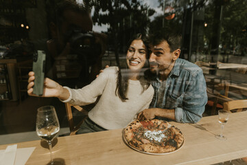 A joyful couple enjoying pizza and capturing a moment together in a cozy cafe on a sunny afternoon