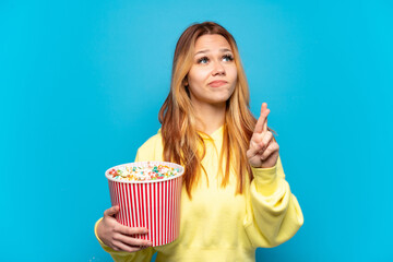 Teenager girl holding popcorns over isolated blue background with fingers crossing and wishing the...