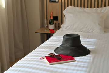 A black sunhat, sunglasses, and a travel journal on a bed with a bottle of champagne in the background, suggesting a luxury vacation.