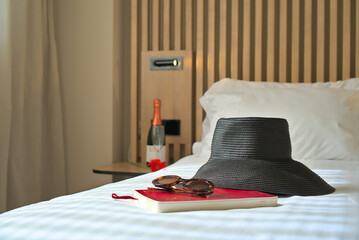 A black sunhat, sunglasses, and a travel journal on a bed with a bottle of champagne in the background, suggesting a luxury vacation.