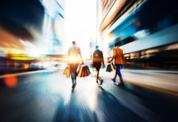 Three people in business attire walk down the street, carrying shopping bags.  The image is blurred and the people's faces are out of focus, they are walking with a purposeful stride.