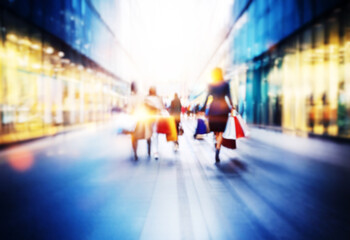 Blurred image of people walking with shopping bags in a city street lined with large glass storefront windows.