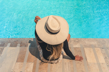 A woman wearing a straw hat sits on a stone staircase by a pool